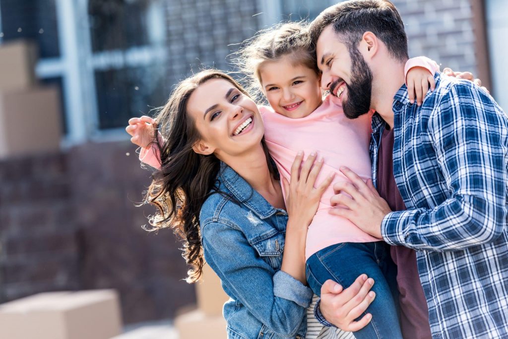 A smiling woman and man hold a young girl between them outdoors, with cardboard boxes blurred in the background.