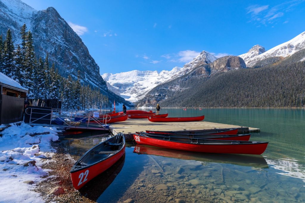 Red canoes docked on a clear lake surrounded by snowy mountains and pine trees under a blue sky.