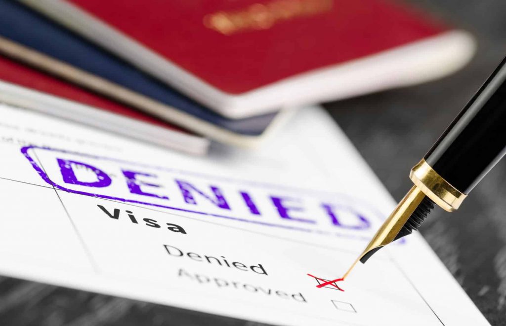 A pen marks “Denied” on a visa application form, with passports in the background and a large “DENIED” stamp visible.
