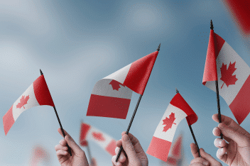 Several hands wave small Canadian flags against a blue sky, each flag featuring a red maple leaf centered between two red bars.