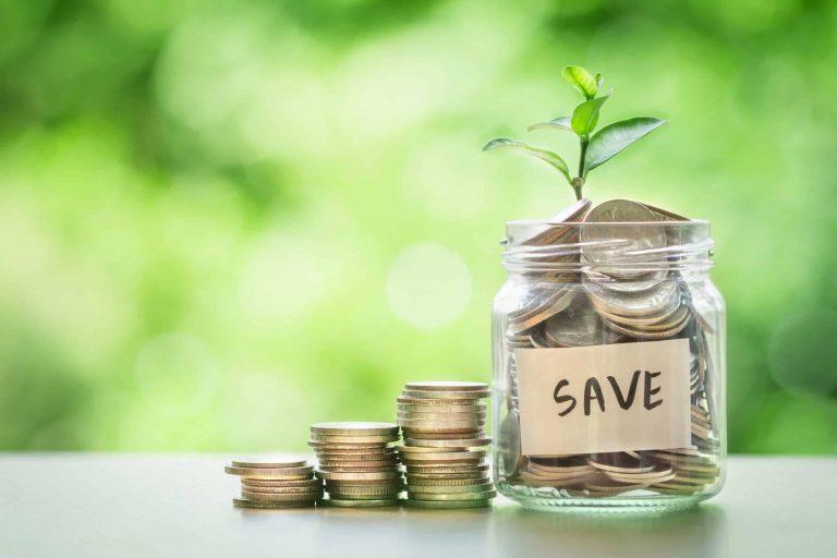A glass jar labeled “SAVE” filled with coins and a small plant sprouting from the top, next to stacked coins, with a green blurred background.
