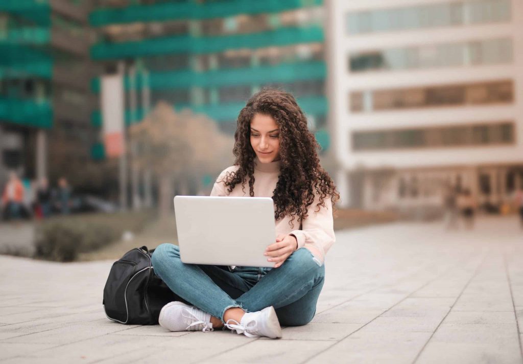 A young woman with curly hair sits cross-legged outdoors, using a laptop on her lap, with a backpack beside her and blurred buildings in the background.