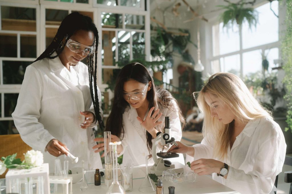 Three women in lab coats and safety goggles conduct experiments with glassware and a microscope in a bright laboratory filled with plants.